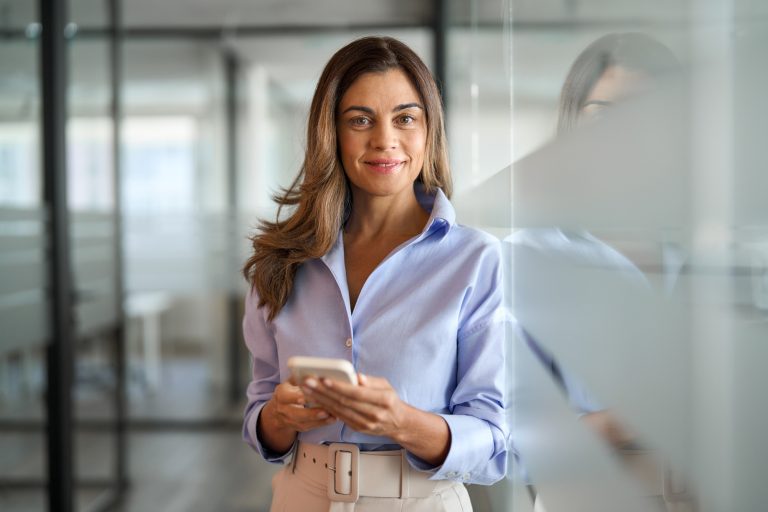Smiling middle aged woman business investor using phone standing in office.